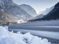 mountains are in the distance behind a snowy lake and road with snow on it and a building in the distance