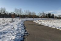 a paved path lined with snow is near the road leading up to a tree covered field