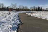 a paved path lined with snow is near the road leading up to a tree covered field