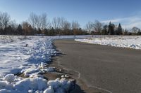 a paved path lined with snow is near the road leading up to a tree covered field