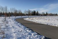 a paved path lined with snow is near the road leading up to a tree covered field