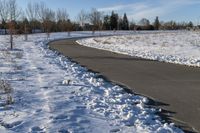 a paved path lined with snow is near the road leading up to a tree covered field