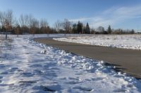 a paved path lined with snow is near the road leading up to a tree covered field