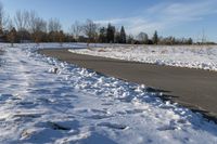 a paved path lined with snow is near the road leading up to a tree covered field