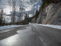 the road is lined by rocks and snow in the distance are trees, and mountains