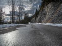 the road is lined by rocks and snow in the distance are trees, and mountains