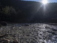 the sun shines brightly behind the trees in the mountain range as a rocky stream reflects the sunlight