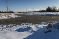 a snowy field with a paved street in the foreground and bare trees in the distance