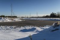 a snowy field with a paved street in the foreground and bare trees in the distance