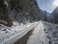 there is snow on the road that goes from the mountains to the trees along with the sun shining through the clouds