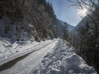 there is snow on the road that goes from the mountains to the trees along with the sun shining through the clouds