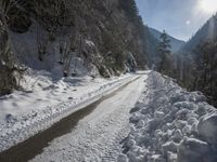 there is snow on the road that goes from the mountains to the trees along with the sun shining through the clouds