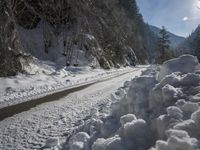 there is snow on the road that goes from the mountains to the trees along with the sun shining through the clouds