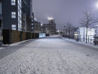 a snowy path near a tall building at night with lights on in the distance and trees