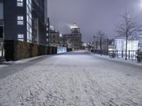 a snowy path near a tall building at night with lights on in the distance and trees