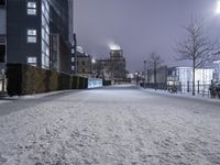 a snowy path near a tall building at night with lights on in the distance and trees