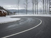 the curve on the side of a snow covered road near trees and water with a mountain in the background