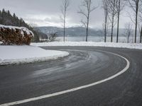 the curve on the side of a snow covered road near trees and water with a mountain in the background