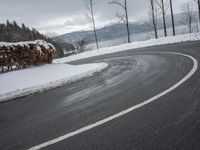 the curve on the side of a snow covered road near trees and water with a mountain in the background