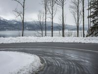 the curve on the side of a snow covered road near trees and water with a mountain in the background