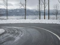 the curve on the side of a snow covered road near trees and water with a mountain in the background