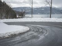 the curve on the side of a snow covered road near trees and water with a mountain in the background