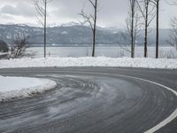 the curve on the side of a snow covered road near trees and water with a mountain in the background