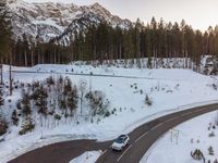 the road with a car that is driving on it in the snow by mountains, and trees and snow covered ground