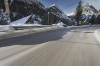 snow on the road and small buildings in the mountains side by side along a path with light poles