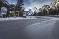 snow on the road and small buildings in the mountains side by side along a path with light poles
