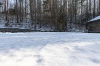 a snow covered yard next to the woods with a bench in it and a building in the background