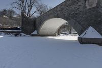 a person walking through snow past an arched bridge over water and buildings on a snowy day