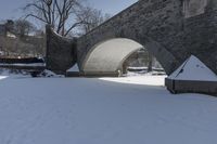 a person walking through snow past an arched bridge over water and buildings on a snowy day