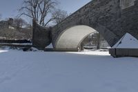 a person walking through snow past an arched bridge over water and buildings on a snowy day