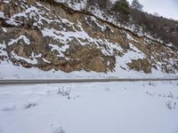 two people are cross country skiing in the snow near a rock wall and road with trees along the side of it