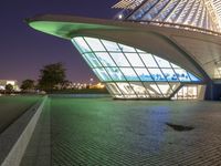 a curved building lit up at night with a large glass window below it and a sidewalk near by