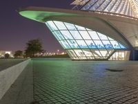 a curved building lit up at night with a large glass window below it and a sidewalk near by