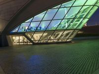 a curved building lit up at night with a large glass window below it and a sidewalk near by