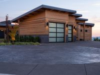 this house has wood clad walls and garage doors along with large windows, and a driveway leading to the front of the house