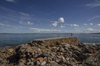 rocks surround the dock on a beach with a lone person standing next to it and looking out at the water