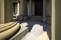 columns are seen outside a stone building near some steps and an entranceway with a wooden door