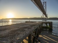 a wooden pier sits in the sand with the sun coming out behind it as it is just under a bridge