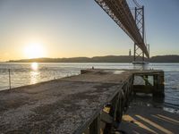 a wooden pier sits in the sand with the sun coming out behind it as it is just under a bridge