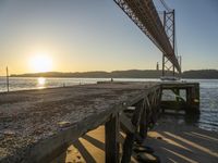 a wooden pier sits in the sand with the sun coming out behind it as it is just under a bridge