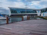 a boardwalk next to boats and a glass building with large windows on the sides and windows on the side