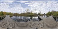 fisheye image of wooden walkway leading into lake and sky with a bridge at bottom