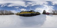 a yellow train passing through the snow near trees and buildings in the background as seen from under a fisheye lens