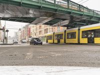 the yellow commuter train is at a stop near cars and buildings in the snow,