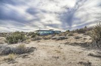 a house sits in a desert like area under a cloudy sky with sparse brush and sparse weeds