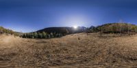 a large grassy hillside with trees and some sun shining above it under blue skies, in the midst of fall